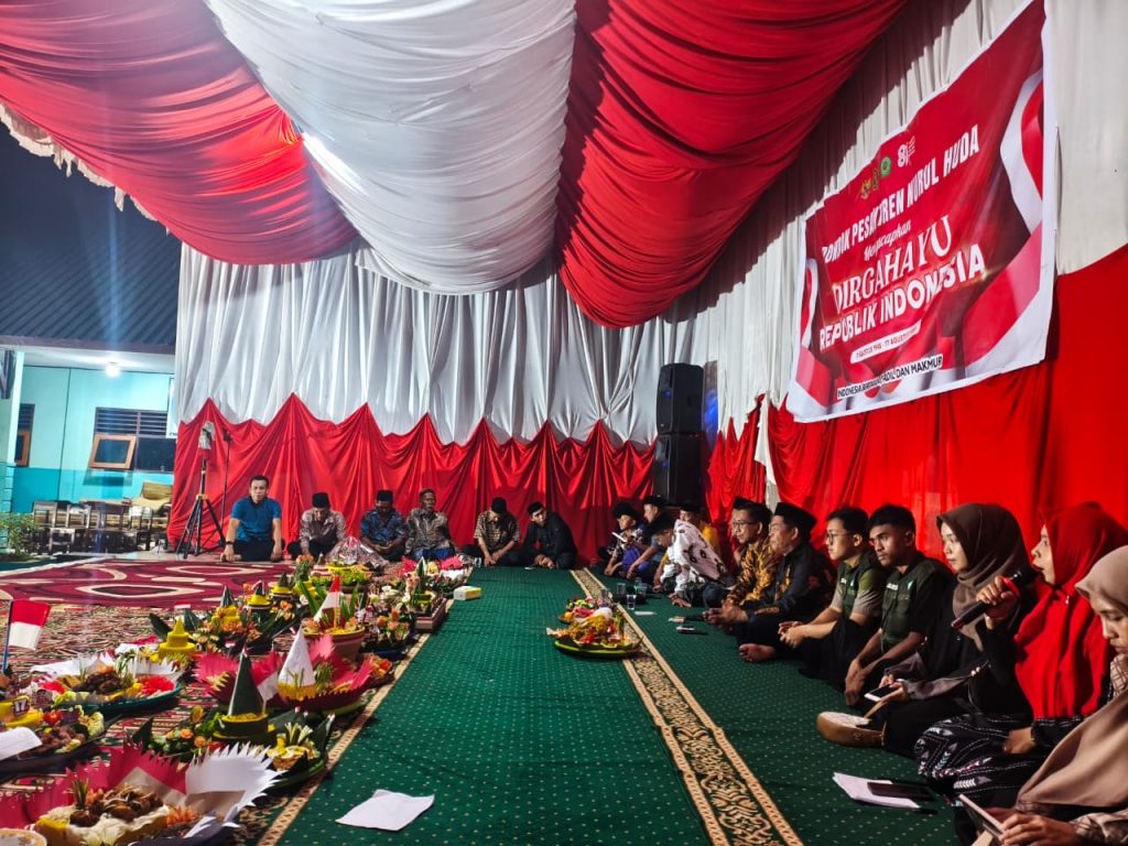 Foto dokumentasi acara malam Dzikir Akbar memperingati HUT RI di Pondok Pesantren Nurul Huda. Tampak panggung megah dihiasi dekorasi kain bernuansa merah dan putih. Di sebelah kanan spanduk besar bertuliskan "DIRGAHAYU REPUBLIK INDONESIA". Di bagian tengah ruangan, deretan tumpeng hias tersaji di atas karpet. Para peserta, termasuk mahasiswa KKA Posko 8 yang mengenakan jas almamater serta para tokoh pesantren dan santri, duduk bersila secara khidmat mengelilingi ruangan.